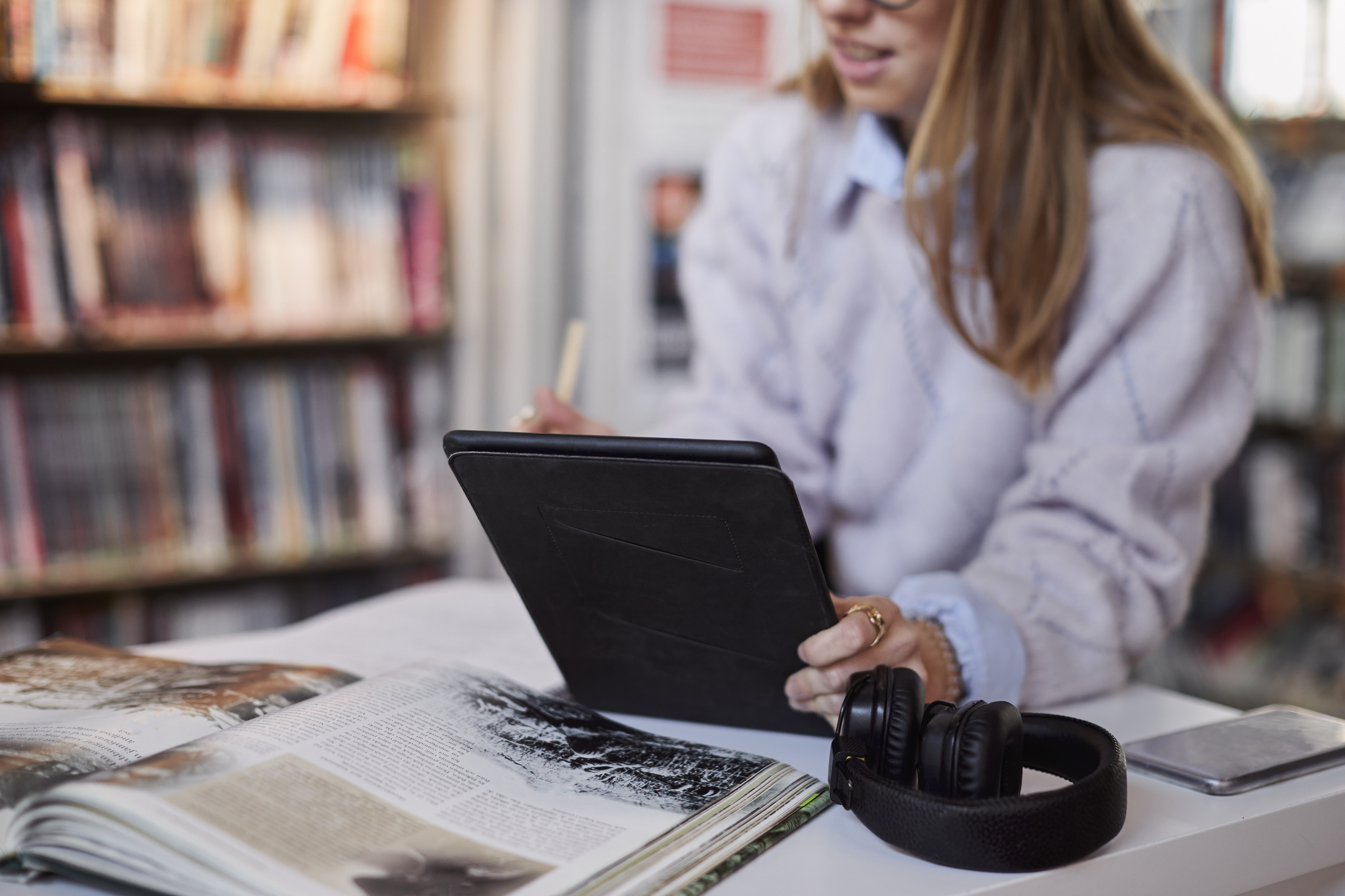 Elev på bibliotek med bok, surfplatta och hörlurar